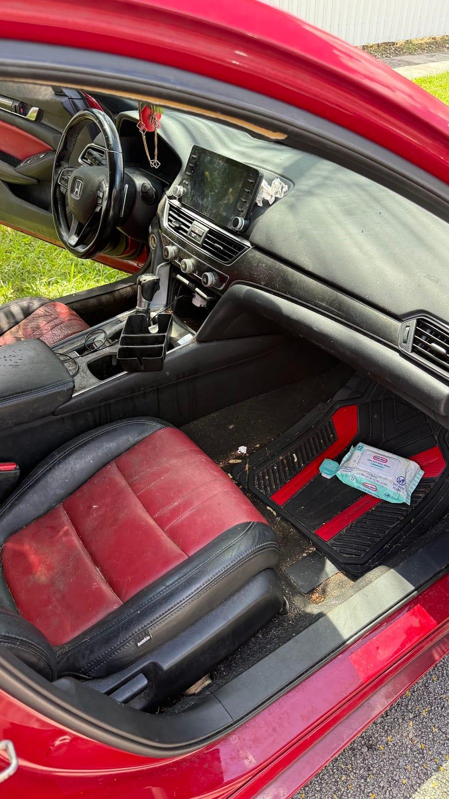 Front passenger area of a red Honda sedan before detailing, with sand and debris across the floor mat and carpet, a discarded wipes packet on the mat, dusty dash and screen, and soiled red-and-black leather seats.